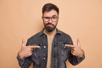 Confident serious bearded adult man makes choose me gesture indicates with index fingers at himeself feels self assured wears glasses black denim jacket poses indoor. Self promoting concept.