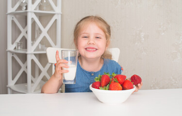 Happy child eating strawberries and drinking milk laughing in the home kitchen. Little smiling girl eats strawberry. Summer time.