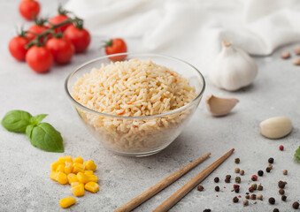 Glass bowl with boiled long grain basmati rice with vegetables on light table background with sticks and tomatoes with corn, garlic and basil.