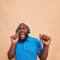 Optimistic joyful dark skinned man dances carefree catches every bit of music has fun listens music via headphones raises arms wears casual blue t shirt isolated over beige background copy space