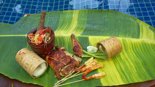 Sticky rice, papaya salad, grilled chicken and grilled shrimp served on a spotted banana leaf poolside..red yellow green speckled banana leaves in the background. food and restaurant concept.