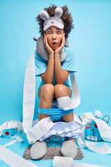 Shocked curly haired African American woman sits on toilet bowl wrapped with paper wears neck...
