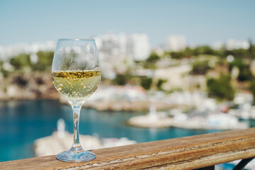 Glass of white wine against the backdrop of the Mediterranean sea and the port with yachts in a tourist town in the summer under sunlight. Summer, travel, lifestyle, relaxation, and enjoyment concept.