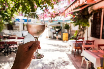 Glass of wine in hand. A glass of young fresh rose wine against the backdrop of a summer cafe in a Mediterranean seaside tourist town in the summer under sunlight. Summer, travel, lifestyle