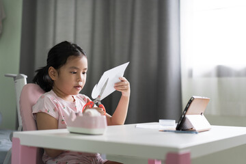 asian child student or kid girl using scissors to cut paper and idea art activity with video call on computer tablet to learning cutting or people enjoy learn from home on smartphone for study online