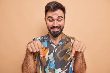 Handsome cheerful bearded man points down gives advice shows link wears colorful shirt has glad...