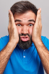 Close up shot of bearded Eurpean man squeezes cheeks makes funny carefree face wears blue t shirt folds lips poses indoor alone against grey studio background. Human facial expressions concept