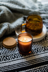 Cozy burning candle in brown glass jar, winter home interior decor with cup and blanket