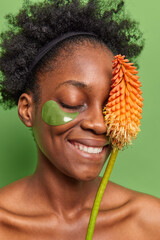 Vertical shot of happy dark skinned female model applies hydrogel patches under eyes to reduce wrinkles holds plant over face uses natural skin care products stands shirtless has curly hair.