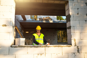 Fototapeta premium A construction engineer and designer in a yellow hardhat studies a drawing of a building on a construction site. A house made of porous concrete blocks, a worker in the process at the window and wall