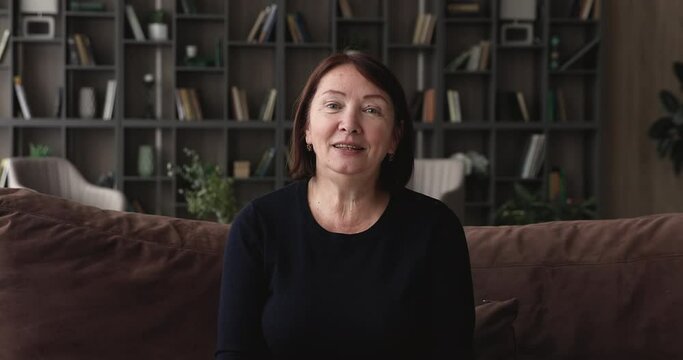 Pleasant beautiful 60s old middle aged woman holding distant web camera conversation with friends, talking remotely with family. Elderly female blogger recording video, sitting on sofa at home.