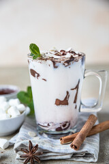 Hot chocolate or cocoa in glass cup with marshmallow, cinnamon and mint, light concrete background.