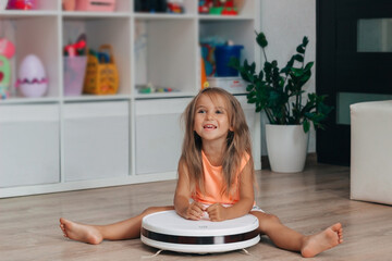  A little girl sits in the children's room on the floor with a robot vacuum cleaner and smiles cheerfully.