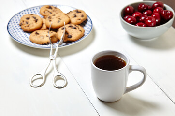 White cup with coffee, traditional cookies and sweet cherries in a ceramic bowl on a white wooden background, closeup. Tasty breakfast, morning routine concept