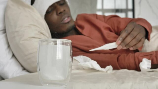 Sad African-American guy with patch on head puts soluble tablet into glass and measures temperature lying in bed close view
