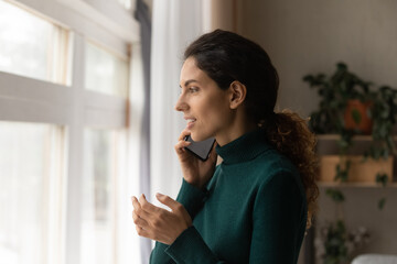 Close up profile view smiling woman taking on phone, standing at home, looking out window, positive young female holding smartphone, speaking, enjoying pleasant conversation with friend, chatting