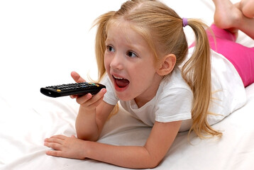 The girl lies on the bed and emotionally watches TV, holding the TV remote control in her hand. The child is watching TV.