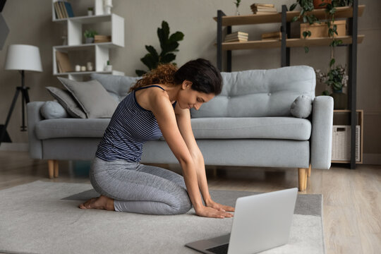 Woman in sportswear stretching at home, involved in online training, sitting on warm floor, motivated young female doing yoga exercises with trainer, morning gymnastics, internet course, class