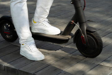 Woman In White Jeans And Sneakers Riding Black Electric Scooter On Wooden Plank Road At Park.