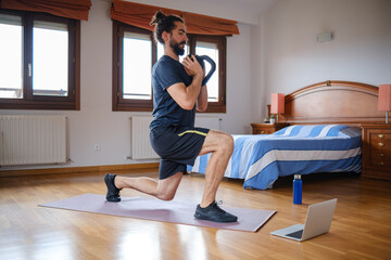 Bearded man doing kettlebell lunges to work out at home while watching an online class on the...