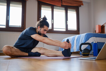 Fototapeta premium Man doing deep stretches at home watching an online exercise class on laptop.