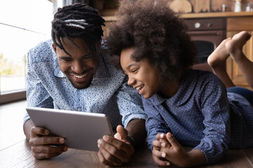 Overjoyed young African American father and teen daughter laugh using modern pad device together. Happy biracial dad and small girl child smile have fun talking on webcam video call on tablet at home.