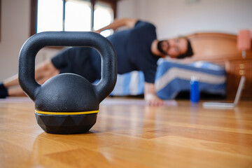 Fototapeta na wymiar Kettlebell in close-up while a man exercises at home with a laptop in the background. Selective focus.