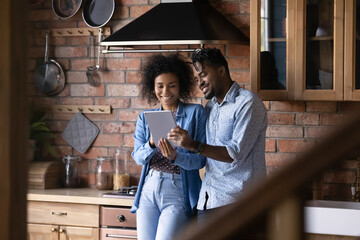 Smiling young African American couple stand in renovated home kitchen use modern tablet gadget shop...
