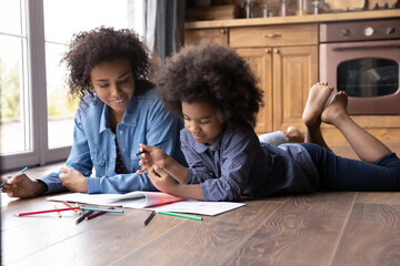 Loving African American mom and small teenage daughter lying on floor in kitchen at home paint draw...