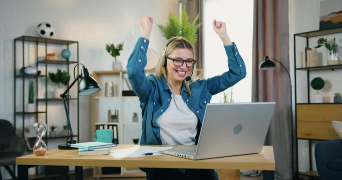 Attractive surprised happy young woman in headset and glasses seeing good news on laptop screen and laughing with raised hands in home office,success and luck concept