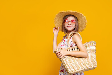 Portrait of happy cheerful girl in summer hat, sunglasses, straw bag over yellow background. 