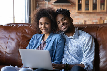 Portrait of smiling millennial African American man and woman sit relax on couch at home using laptop together. Happy biracial couple rest on sofa browse internet communicate online on computer.