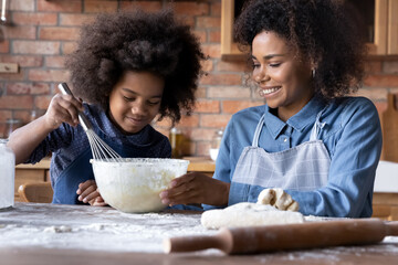 Loving smiling young African American mother teach small teen daughter cooking making dough...