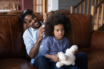 Loving young African American father help brush hair of small teen daughter relax at home together. Happy ethnic dad take care of cute little teenage girl child on family weekend. Parenthood concept.