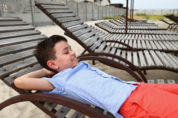 Happy teenage boy lying down on a wooden chaise lounge. Handsome child resting on the deck chair during summer country vacations.