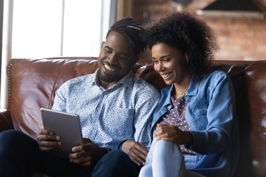 Smiling young African American couple rest talk speak on webcam video call on tablet gadget together. Happy ethnic man and woman sit on sofa have virtual event on pad. Communication concept.