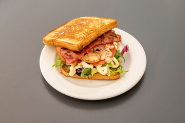 Grilled buttered sliced bread sandwich with roast chicken, fried bacon, sliced tomato, lettuce and mayonnaise on white plate