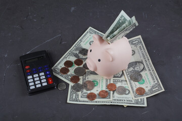 pig piggy bank stands on one-dollar bills next to a calculator on a dark concrete background.