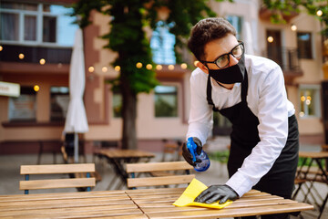 Disinfecting cafe tables to prevent COVID-19. Young waiter wearing protective face mask while cleaning tables while working in a cafe.