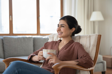 Smiling young ethnic female renter sit rest in armchair drink hot coffee or tea look in distance...