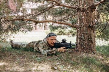 Man is lying on the green grass and looking into the optical sight a weapon. People playing laser tag shooting game in outdoor. War simulation game