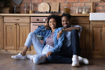 Happy millennial African American couple sit on wooden floor in cozy renovated kitchen moving in together. Smiling young ethnic man and woman spouses relax at home relocate to own house. Rent concept.
