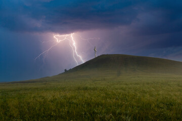 Summer awe landscape with flashes of bright lightning on dark cloudy sky during a thunderstorm over the Orthodox cross at the top of the hill. Drokinskaya mountain near Krasnoyarsk, Russia