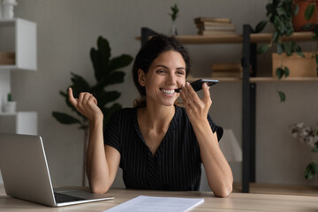 Smiling businesswoman recording audio voice message on smartphone, sitting at desk with laptop,...