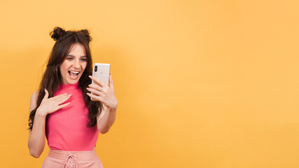 A smiling woman holds a smartphone on which she records a video message or talks in a video chat, standing on a yellow background. A copy of the space. Space for text