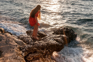 A girl in a red dress on the rocks on the seashore. Sea sunset. Journey.