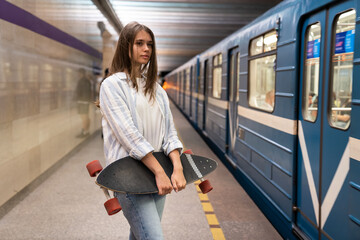 Young girl waiting for subway car at underground metro station alone holding longboard skate....