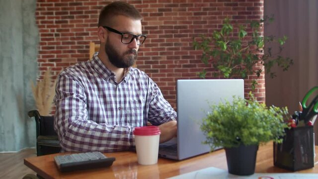 Wooden cubes with inscription disabled, man in wheelchair and glasses looks straight and smiles while typing on laptop. Concept of self acceptance
