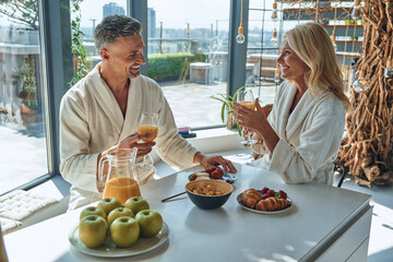 Beautiful mature couple in bathrobes enjoying breakfast together while spending time in the domestic kitchen