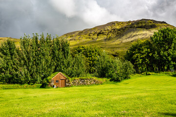 Obraz premium Fresh summer green view from Southern Iceland. Small stone service house covered in moss and grass next to small forest and hill